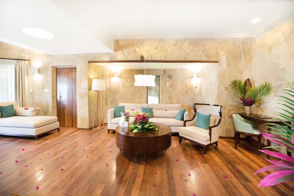 Hotelzimmer mit Holzboden, Steinplatten an den Wänden, cremefarbenem Sofa und rosa Blütenblättern auf dem Boden