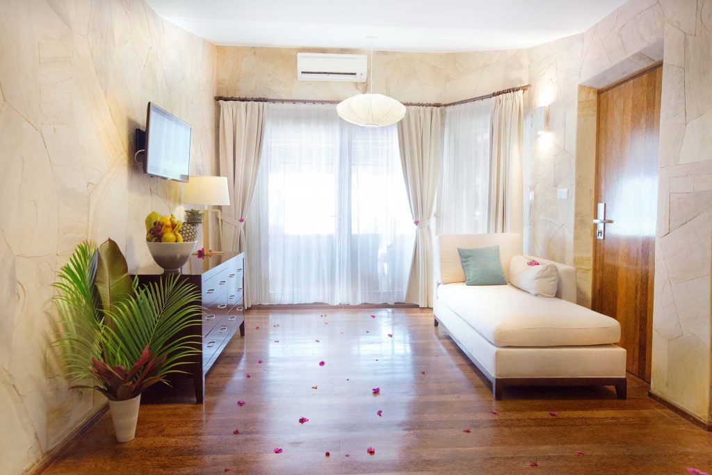 Hotelzimmer mit beiger Chaiselongue, hellem Holz, dunkler Kommode mit Obstschale, Flachbildfernseher und Tageslicht von zwei.