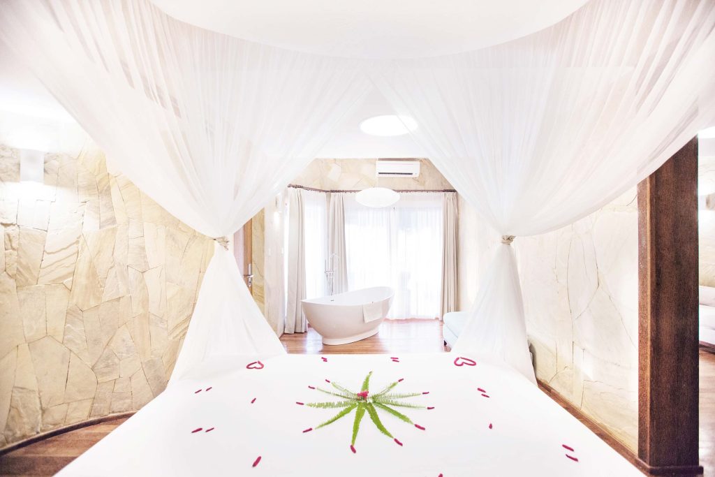 Hotelzimmer mit großem Bett, weißen Vorhängen, freistehender Badewanne vor Fenster und Natursteinwänden