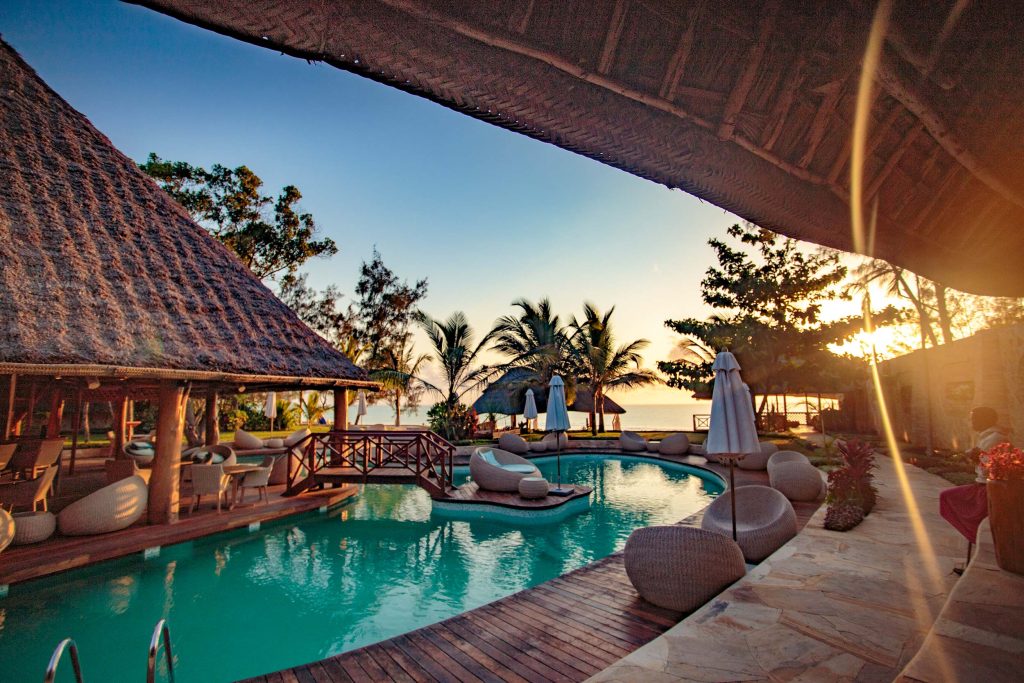 Außenbereich von Tulia Zanzibar mit Swimmingpool, Holzdeck, gepolsterten Liegen und Strohdach-Pavillon bei Tageslicht