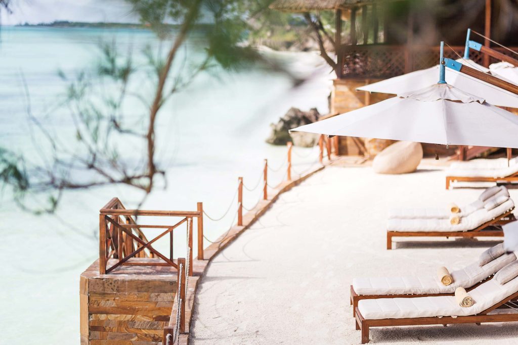 Strandbereich im Tulia Zanzibar mit weißen Sonnenliegen, Sonnenschirmen und Holzweg entlang des Wassers