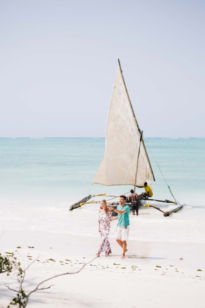 Paar spaziert am weißen Sandstrand mit türkisem Wasser und traditionellem Holzsegelboot in Sansibar