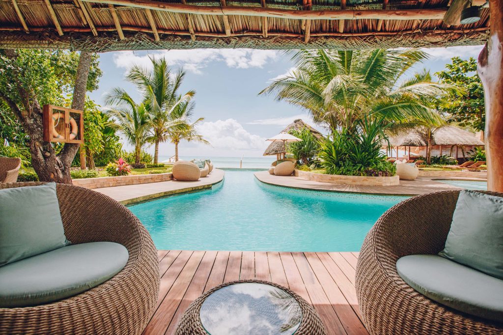 Außenbereich des Tulia Zanzibar Resorts mit Pool, Holzdeck, zwei geflochtenen Sesseln und Palmen im Hintergrund