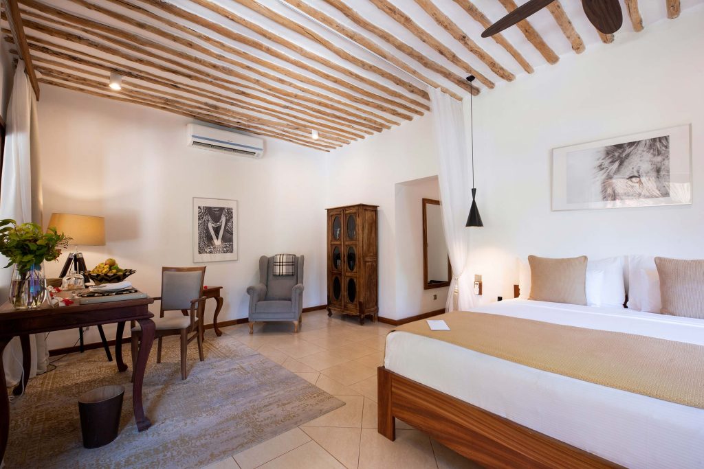Hotelzimmer mit großem Bett, weißer Bettwäsche, Holzbalkendecke, Steinboden und Schreibtisch in Sansibar