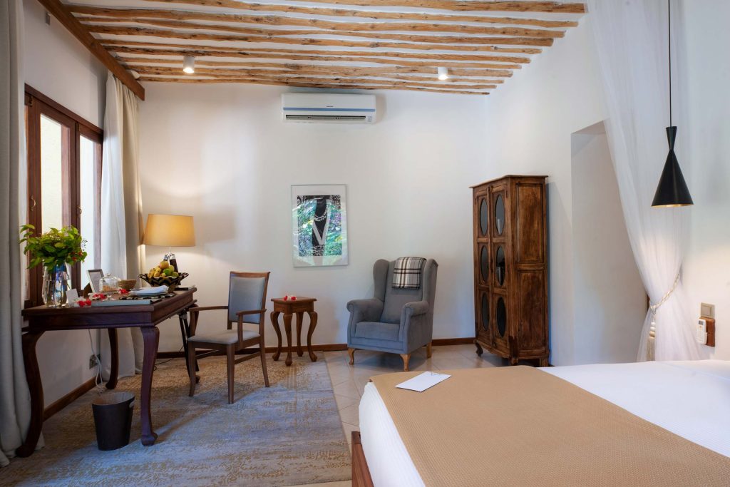 Hotelzimmer mit freiliegenden Holzbalken, Holztisch, grauem Sessel und großen Fenstern in Sansibar