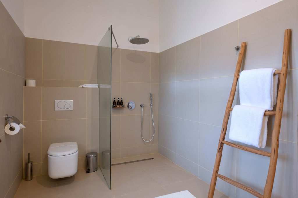 Modernes Badezimmer mit hellbeigen Fliesen, offener Dusche, Holzleiter und weißen Handtüchern in Sansibar