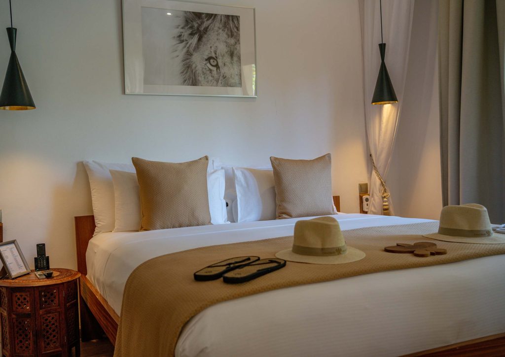 Hotelzimmer mit zentralem Bett, weißer Bettwäsche, beigen Kissen, Holzboden und zwei schwarzen Hängeleuchten