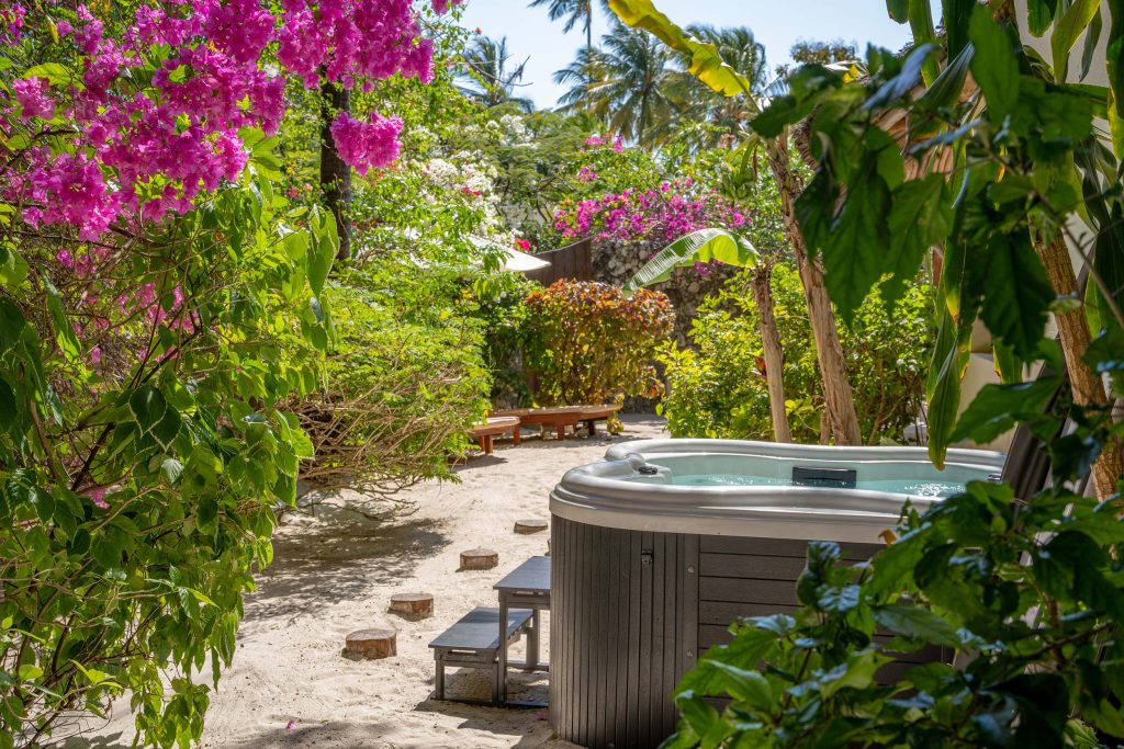 Whirlpool aus Kunststoff und Holz im tropischen Garten mit Sandboden und pinken Bougainvillea, Liegestühle im Hintergrund