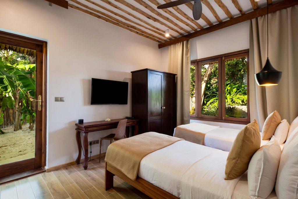 Hotelzimmer mit zwei Einzelbetten, dunklem Holzschrank, Schreibtisch und großen Fenstern mit Blick auf Garten in Sansibar