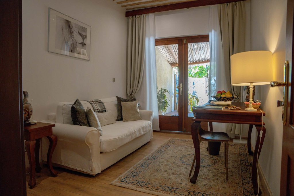 Wohnzimmer mit weißer Couch, gemustertem Teppich, Holztischen, Lampe und Tageslicht durch Glastür