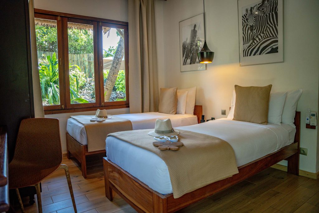 Hotelzimmer mit zwei Einzelbetten in Holzrahmen, weißen Bettlaken, großen Fenstern und Blick auf Grün in Sansibar