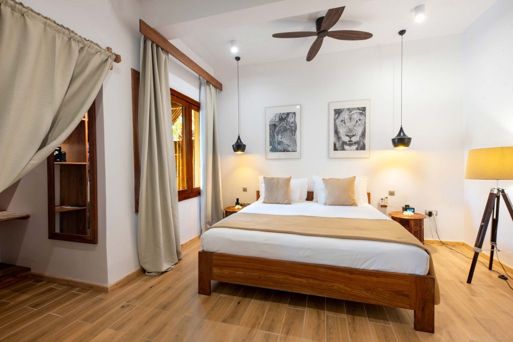 Hotelzimmer mit großem Holzdoppelbett, Holzfliesenboden, Schwarz-Weiß-Tierbildern und modernem Deckenventilator