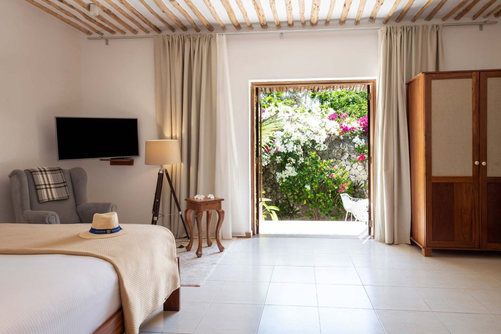 Helles Hotelzimmer mit großem Bett, grauem Sessel, Stehlampe und Blick auf Garten in Sansibar