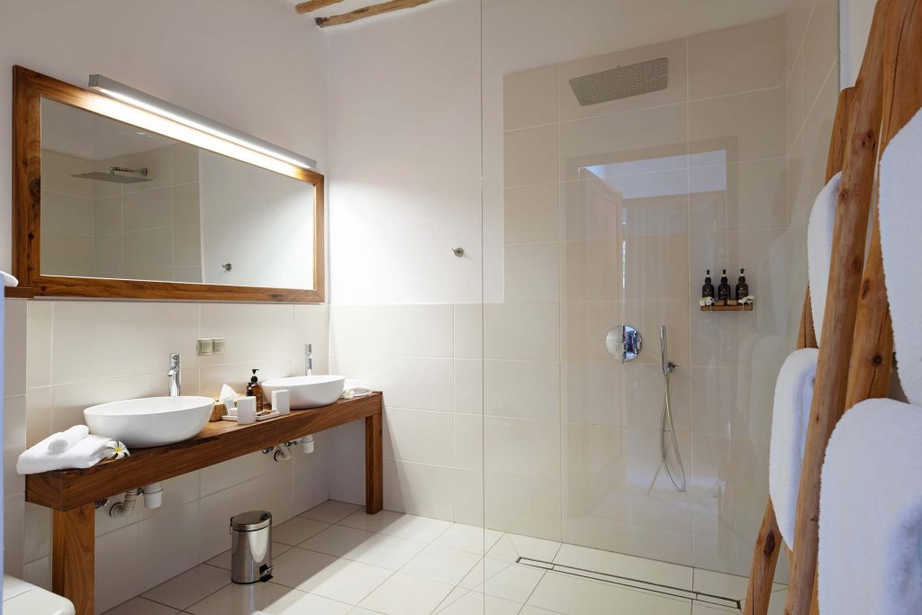Modernes Badezimmer mit dunklem Holzwaschtisch, zwei weißen Waschbecken, großem Spiegel und Glasdusche im Zanzibar White.