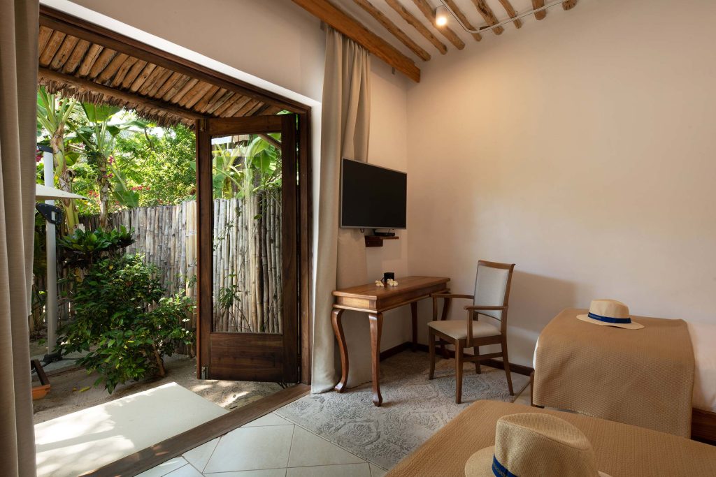 Hotelzimmer mit Holzmöbeln, Schreibtisch, offener Holztür zum Garten mit Bambuszaun und tropischen Pflanzen