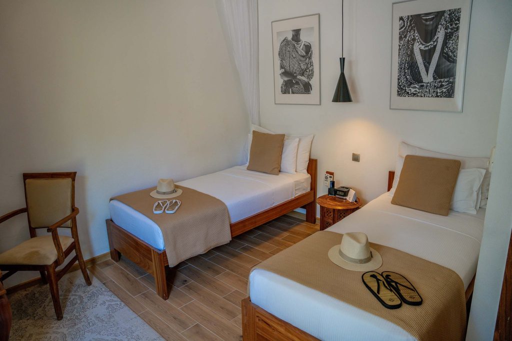 Hotelzimmer mit zwei Einzelbetten, weißen Laken, beigefarbenen Überwürfen, Strohhüten und dunklen Holzmöbeln