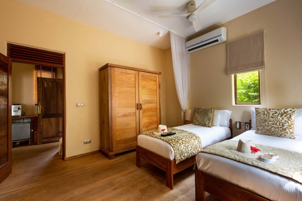 Hotelzimmer mit zwei Einzelbetten aus Holz, weißen Laken, goldgemusterten Kissen, Holzschrank, Holzfußboden
