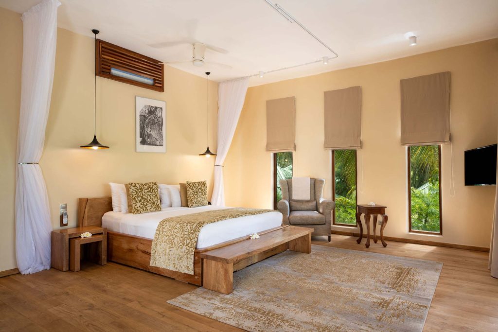 Hotelzimmer mit großem Holz-Himmelbett, grauem Sessel, Holztisch, Holzfußboden und schmalen Fenstern mit Blick auf tropische.