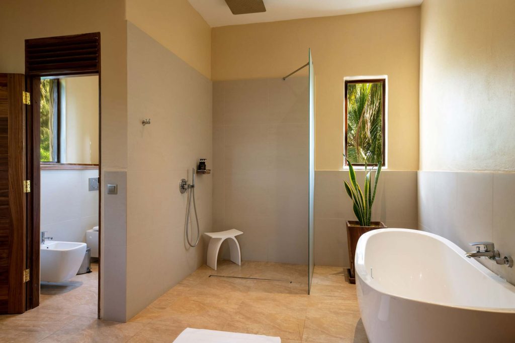 Modernes Hotelbadezimmer mit freistehender weißer Badewanne, begehbarer Dusche, Natursteinboden und Fenster mit Blick.