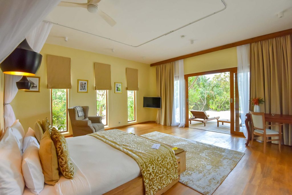 Geräumiges Hotelzimmer mit großem Bett, weißen und goldenen Kissen, Holzboden und geöffneter Terrassentür