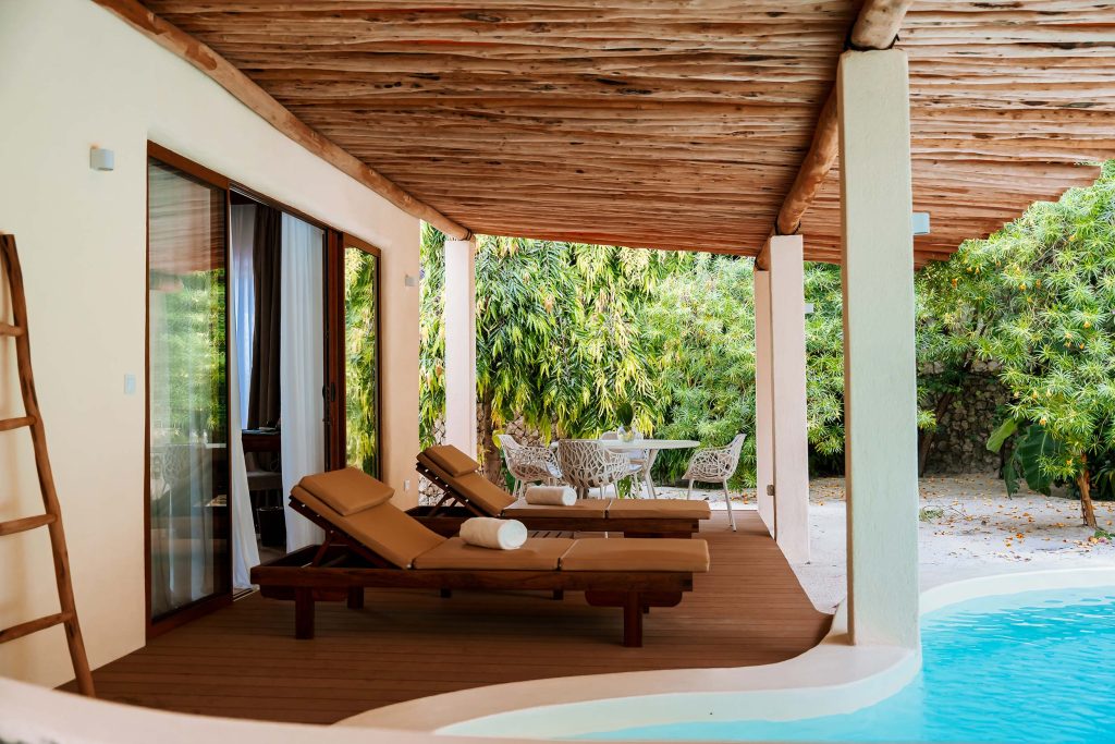 Überdachte Terrasse mit zwei hölzernen Liegestühlen, rundem Metalltisch und Pool im Hintergrund in Sansibar