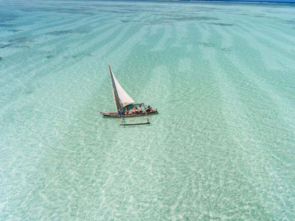 Holzsegelboot mit weißem Segel und drei Personen auf klarem türkisfarbenem Wasser bei Tageslicht