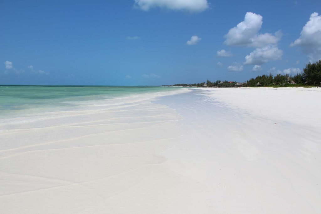 Weiter weißer Sandstrand mit türkis schimmerndem Meer und blauem Himmel mit weißen Wolken bei Sansibar
