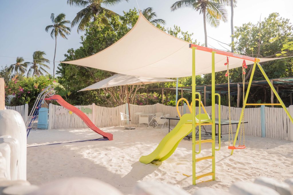 Außenbereich mit Klettergerüsten, gelber und roter Rutsche, Schaukel und weißem Sonnensegel in hellem Sand