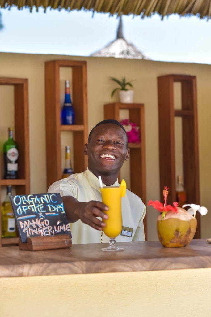 Offene Hotelbar mit hellem Holztresen, Strohdach und Barkeeper mit gelbem Cocktail in Sansibar