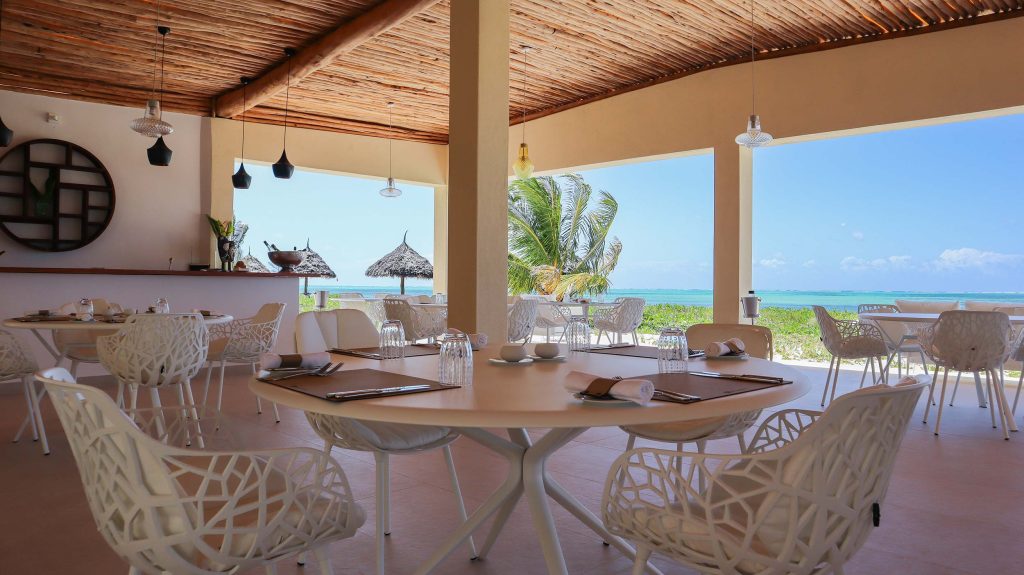 Hotelrestaurant mit runden Tischen, weißen Stühlen, Holzdecke und Blick auf Palmen und türkisfarbenes Meer in Sansibar