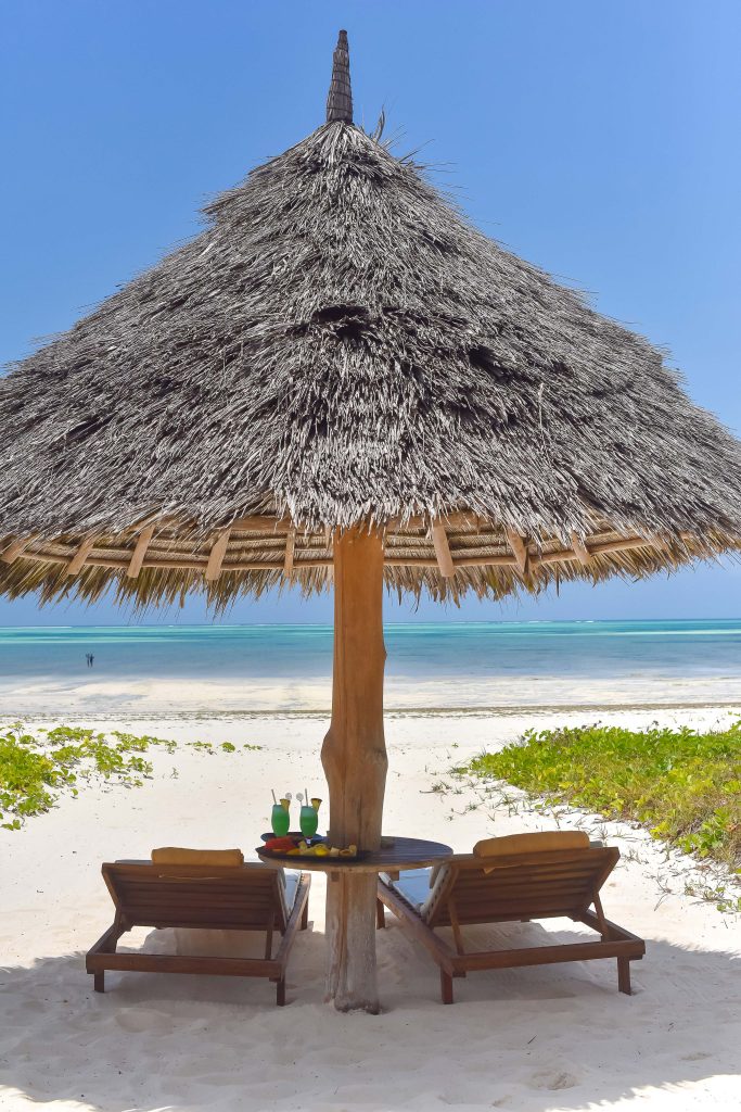 Strand mit Strohdach-Sonnenschirm, zwei Holzliegen, Holztisch mit Getränken, weißer Sand.