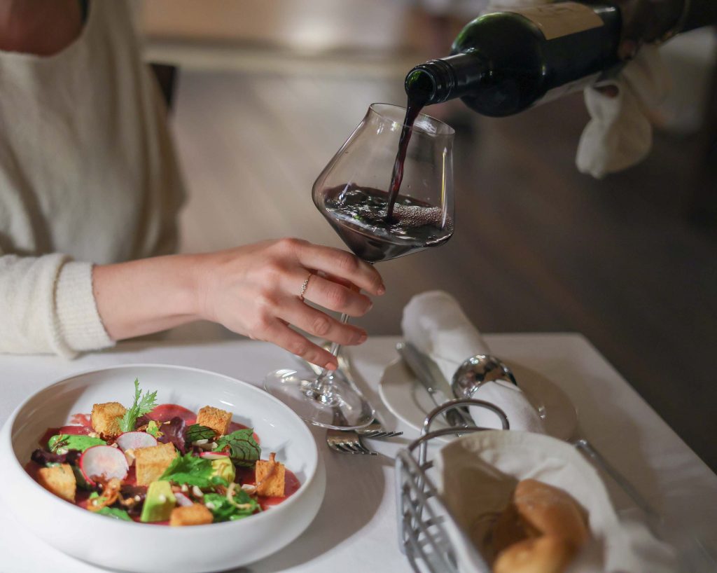 Person gießt Rotwein in ein Weinglas auf weiß gedecktem Tisch mit Salat, Brotkorb und Servietten im Hotelrestaurant