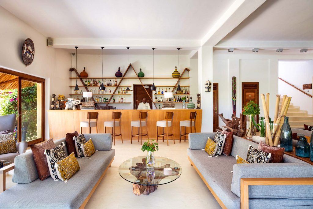 Innenraum der Hotelbar mit grauen Sofas, bunten Kissen, Holztheke, Barhockern und Weinregal in Sansibar