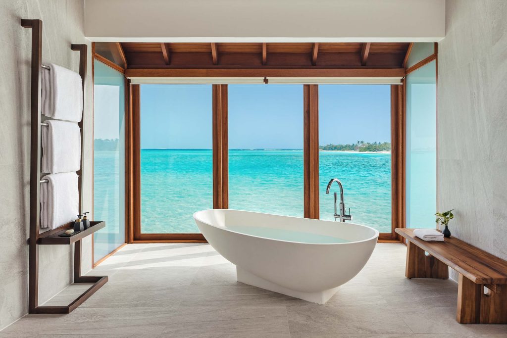 Freistehende weiße Badewanne vor Fensterfront mit Blick auf türkisfarbenes Meer und blauen Himmel