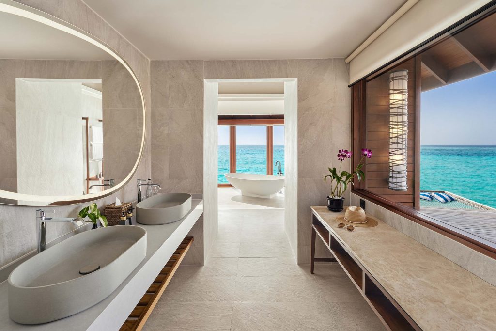 Badezimmer mit zwei grauen Steinwaschbecken, beigen Fliesen und freistehender Badewanne vor Meerblick