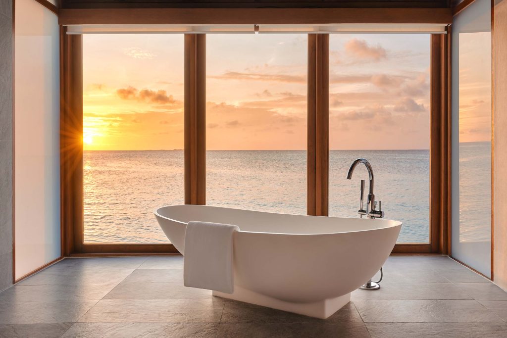Badezimmer mit freistehender weißer Badewanne vor bodentiefen Holzfenstern mit Meerblick bei Sonnenuntergang