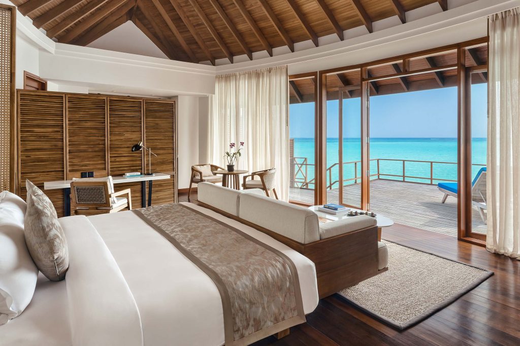Hotelzimmer mit großem Bett, Holzdecke, Schreibtisch und Glasfenstern mit Blick auf Terrasse und Meer