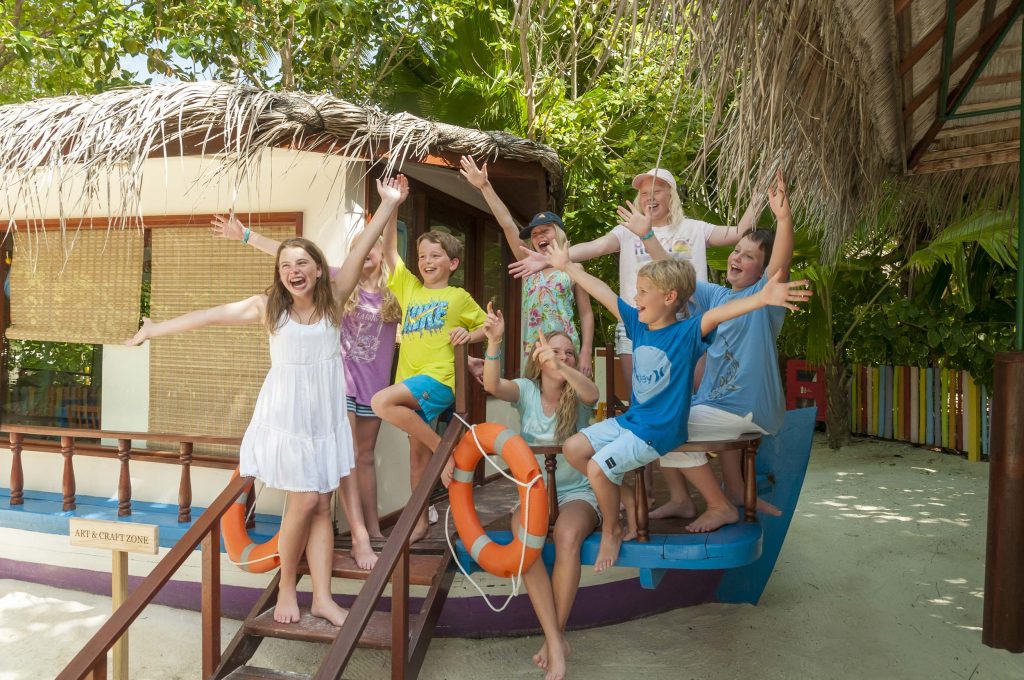 Gruppe von Kindern spielt auf einem bunten Holzboot aus Blau und Lila vor einer Palmwedel-Hütte und grünem Laub
