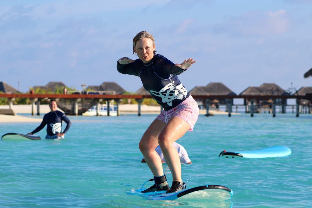 Person beim Surfen auf klarem türkisfarbenem Meer vor Stelzenbungalows aus Holz bei blauem Himmel