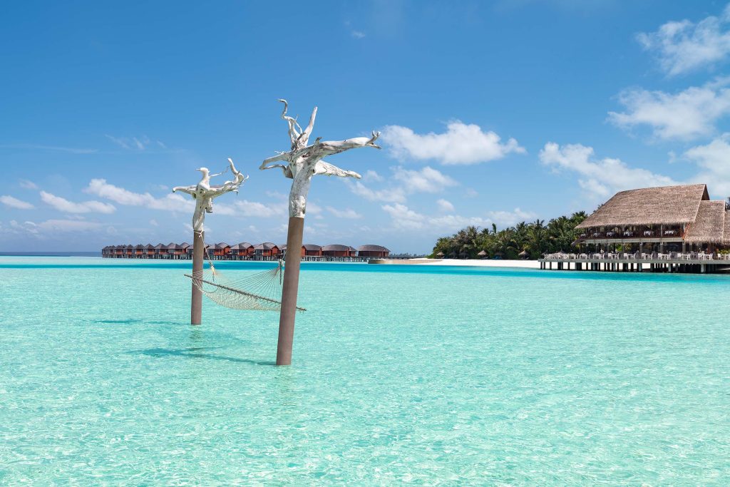 Hängematte zwischen Holzmasten über türkisfarbenem Wasser mit Wasserbungalows und Naturdach im Hintergrund