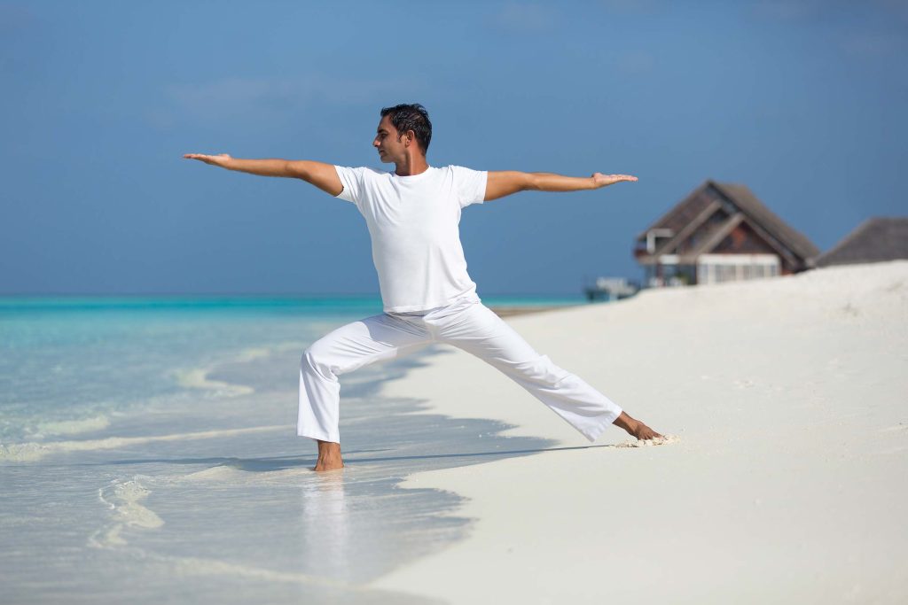 Mann in Yoga-Pose steht im flachen Wasser am Strand mit hellem Sand und türkisfarbenem Meer, Strohhütten im Hintergrund