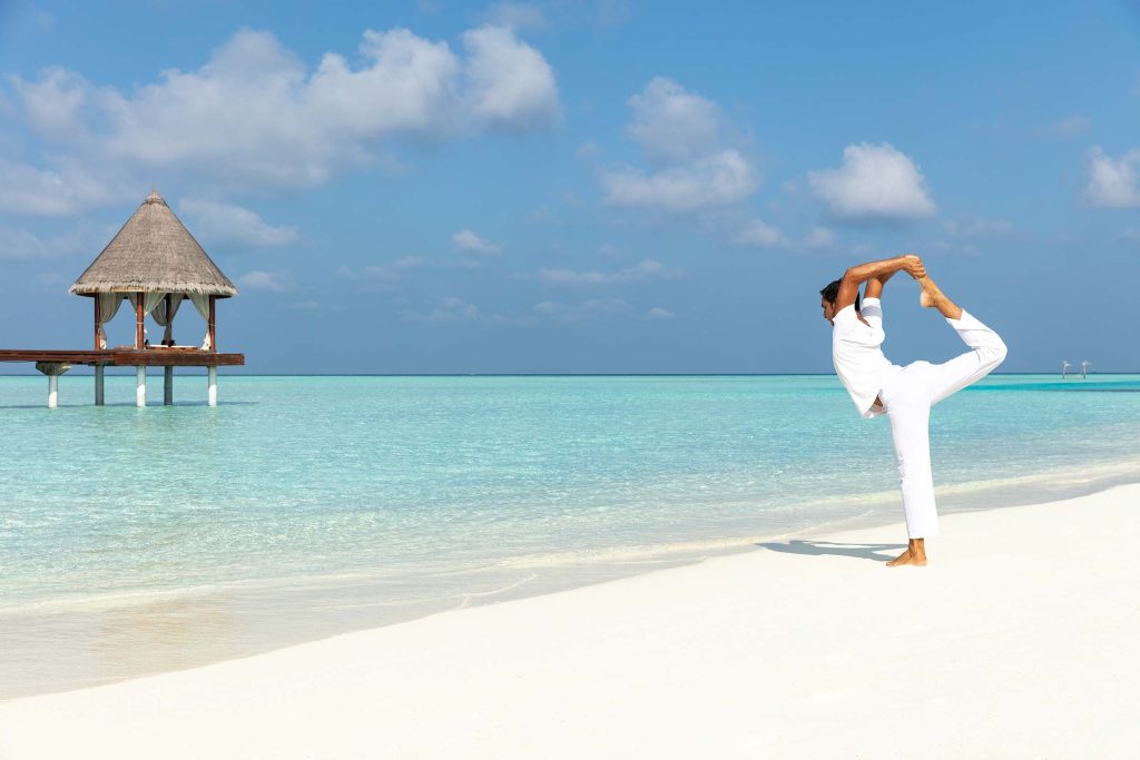 Strand mit weißem Sand, türkisfarbenem Wasser, hölzerner Plattform mit Strohdach und Person in weißer Kleidung beim Yoga