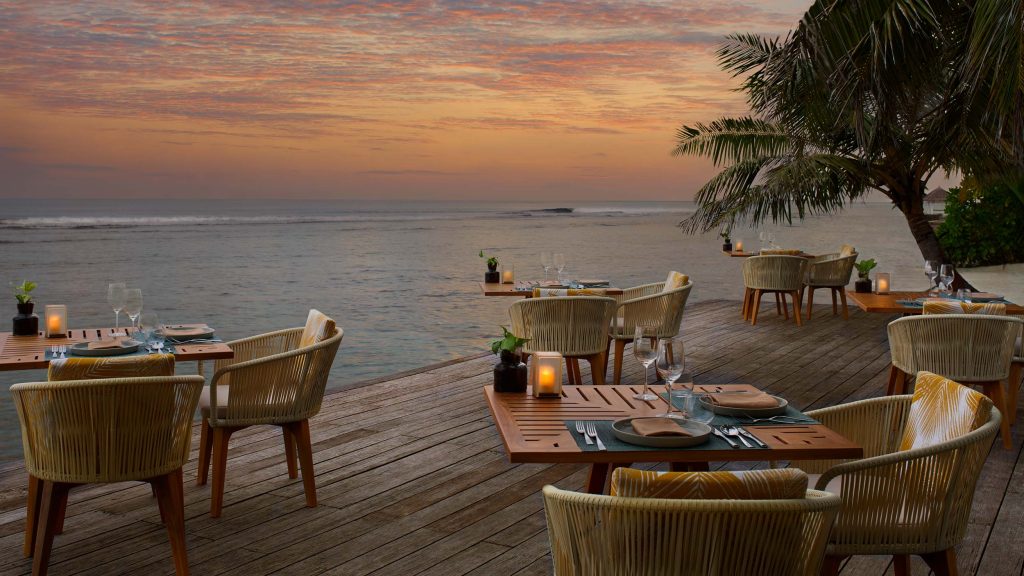 Außenrestaurant mit Holztischen und gepolsterten Stühlen auf Holzterrasse am Meer bei Sonnenuntergang