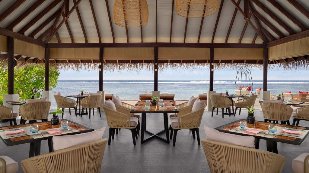 Offener Restaurantbereich mit hellen Rattanmöbeln und Holztischen, Blick auf das blaue Meer und tropische Pflanzen