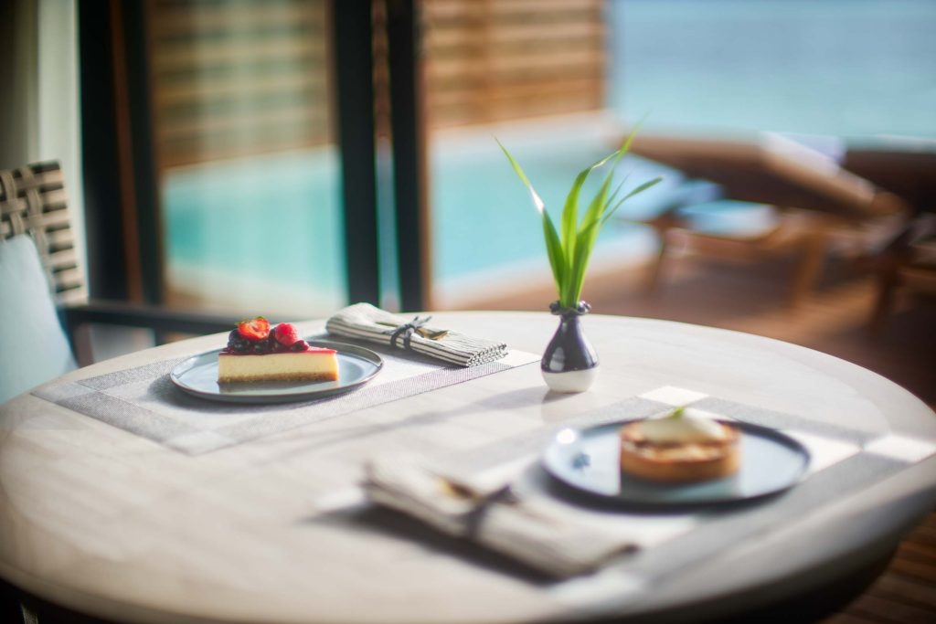 Runder Holztisch mit zwei Desserttellern, Stoffservietten und Pflanze, Terrasse und Wasser im Hintergrund