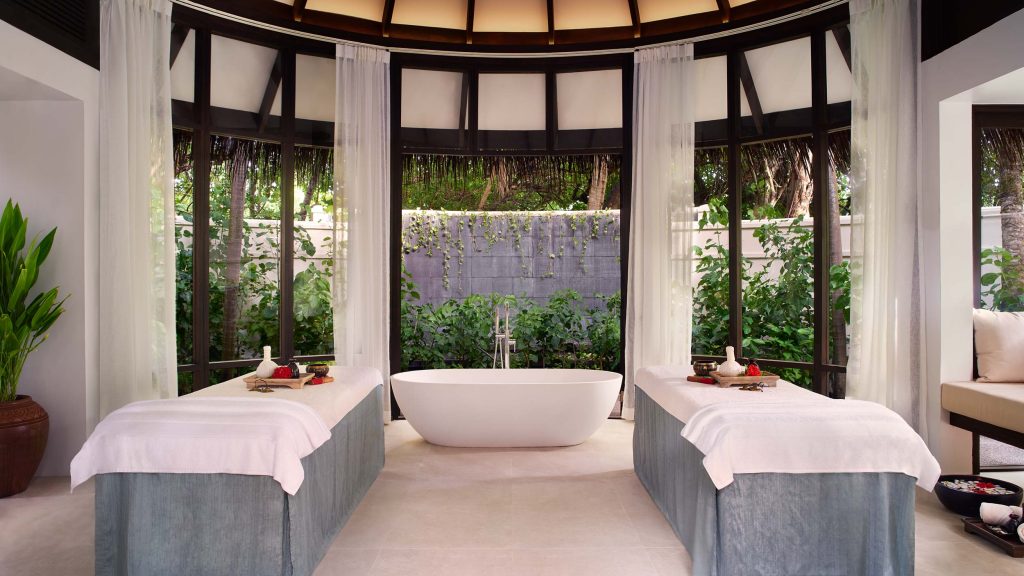 Spa-Raum mit weißer freistehender Badewanne, zwei Massageliegen, großen Fenstern und grünem Pflanzenblick