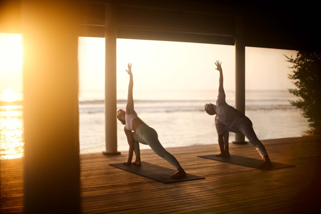 Zwei Personen praktizieren Yoga auf einer Holzterrasse mit weißen Säulen und Meerblick bei Sonnenuntergang