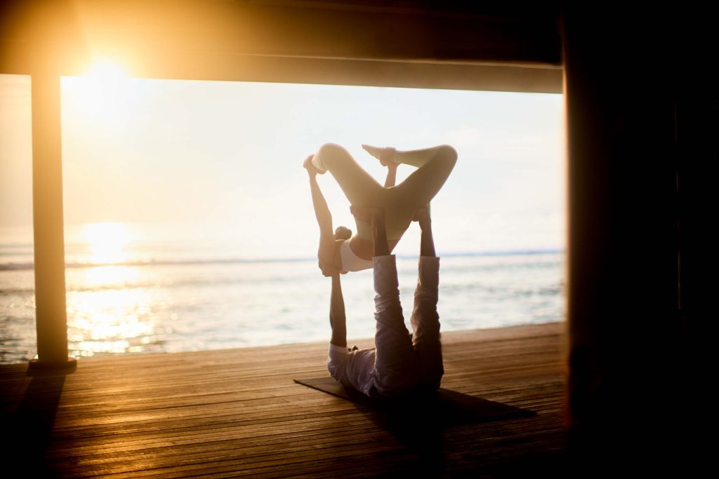 Zwei Personen praktizieren Yoga auf einer dunklen Holzterrasse mit Meerblick bei Sonnenuntergang