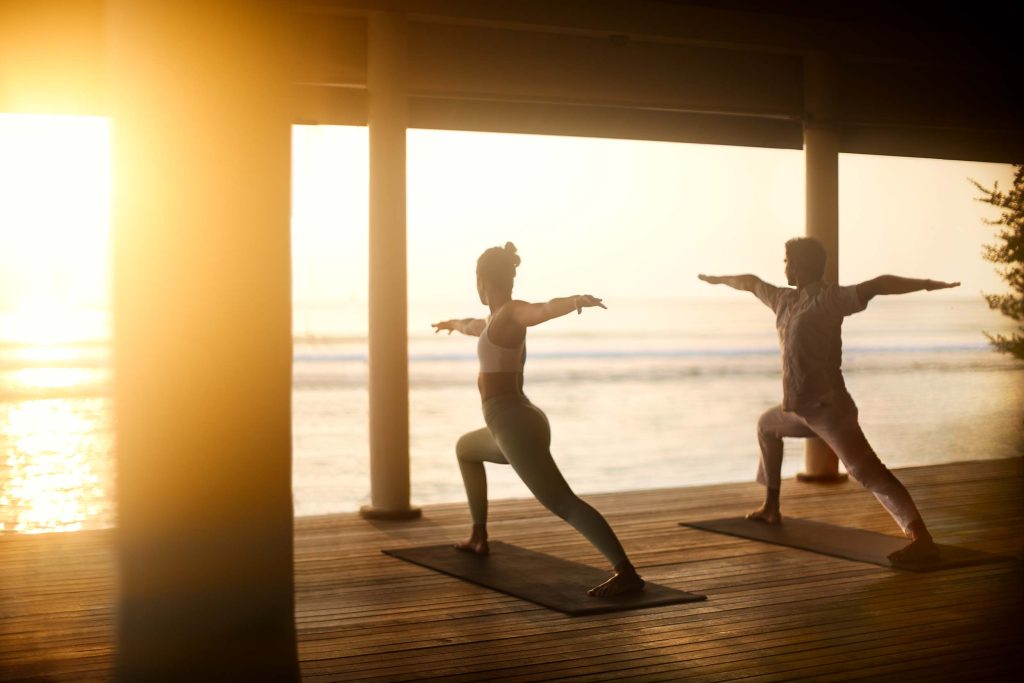 Zwei Personen in Yoga-Posen auf Holzplanken in offenem Raum mit Säulen und Meerblick bei Sonnenuntergang