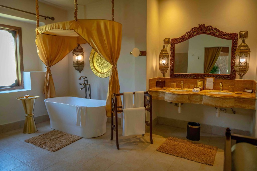 Badezimmer mit freistehender weißer Badewanne unter goldfarbenem Baldachin, Fenster und Marmorwaschbereich